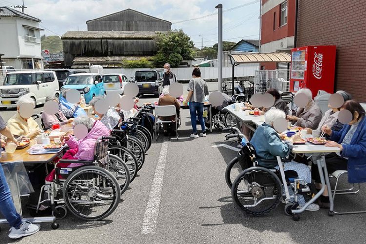 屋外駐車場での入居者による食事・交流イベント。車いすユーザーも参加しており、レクリエーション活動の様子が伺える。